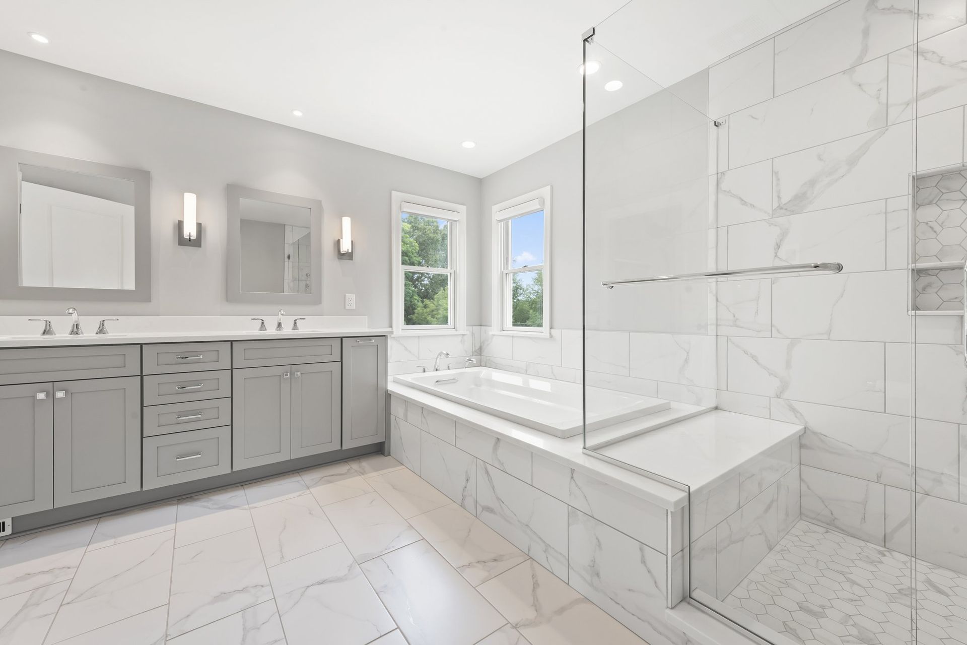 Primary bathroom with a gray double vanity, a tub with a tiled deck, and a frameless glass shower in marble-look tile.