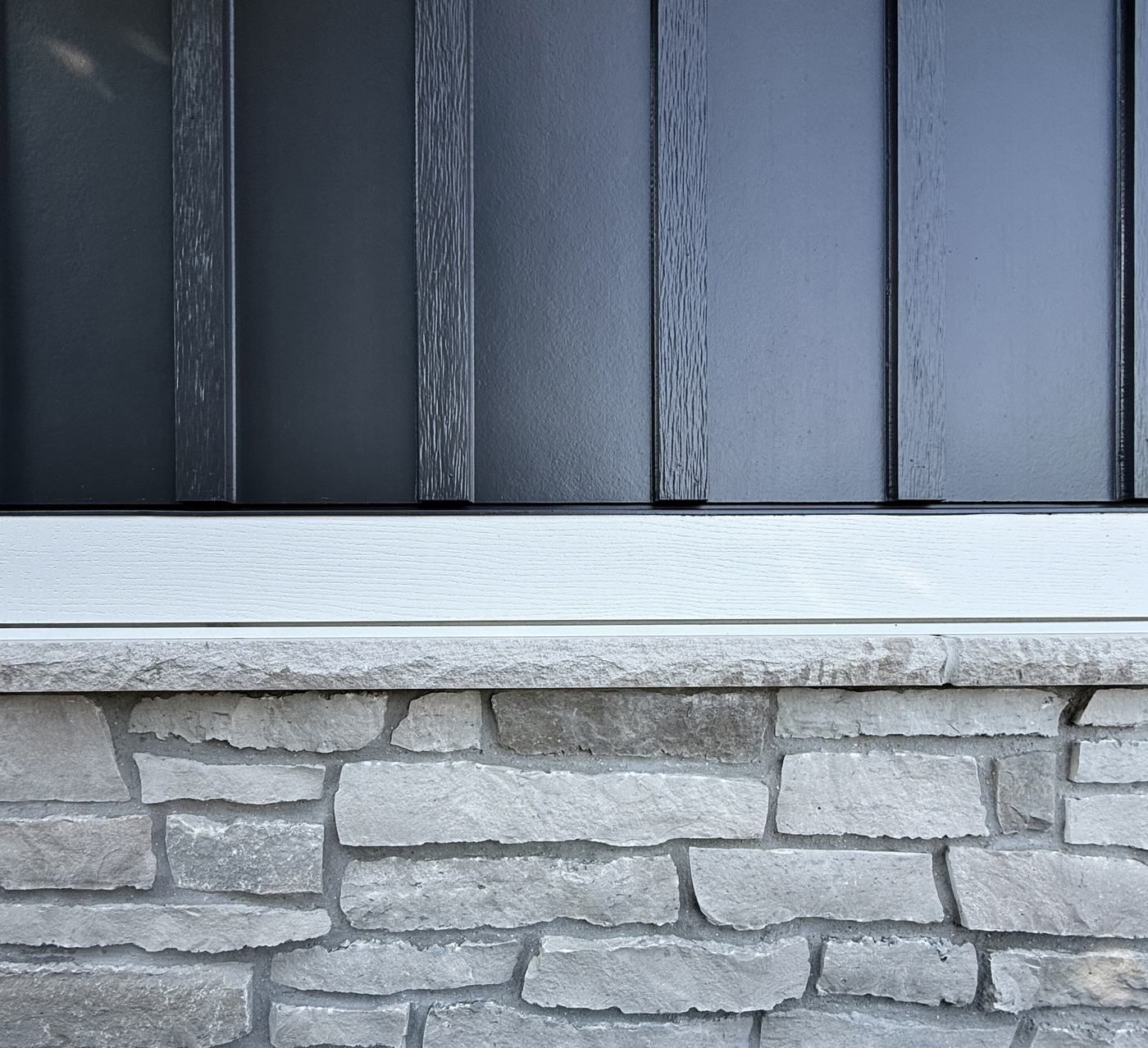 Detail of dark board and batten siding over a white trim band, a stone sill, and natural stone.