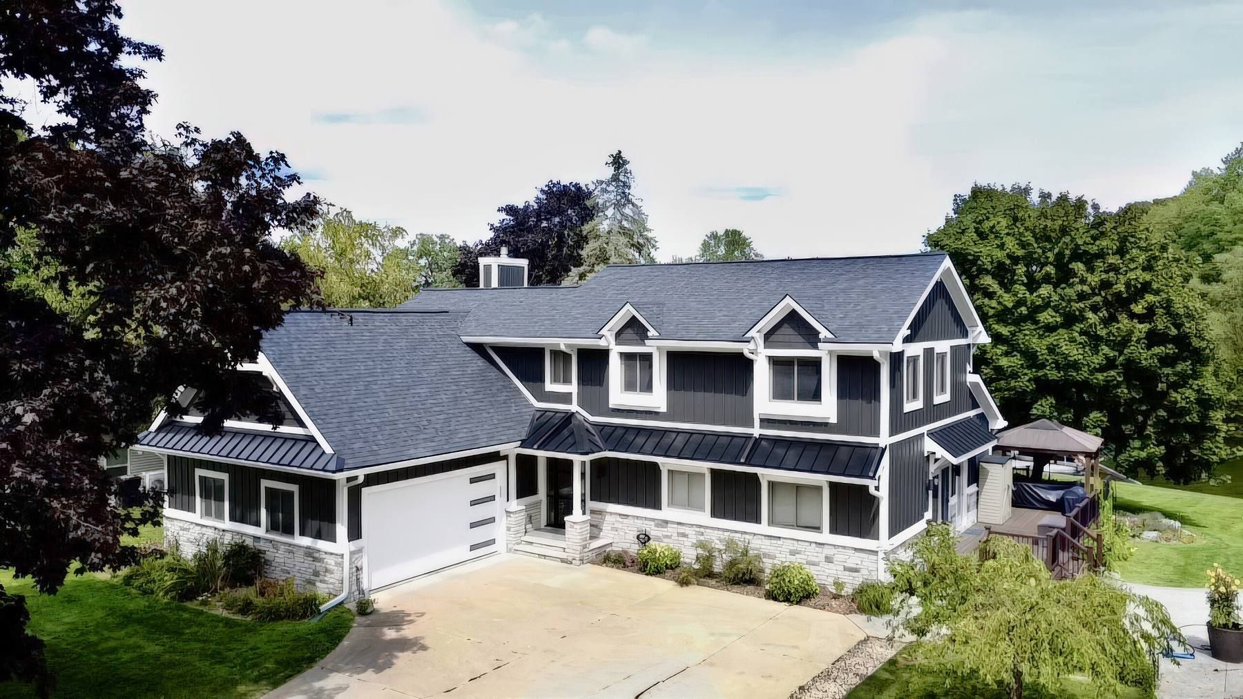Aerial view of the remodeled house with dark board and batten siding, white trim, stone along the base, metal eyebrow roofs, and a new front porch.