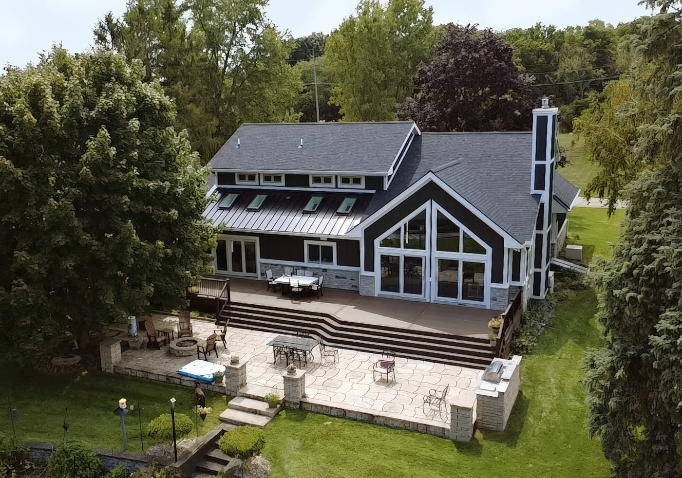 Aerial view of the lake side of the house with a tall glass gable, standing seam metal lower roofs, skylights, and the deck and patio.