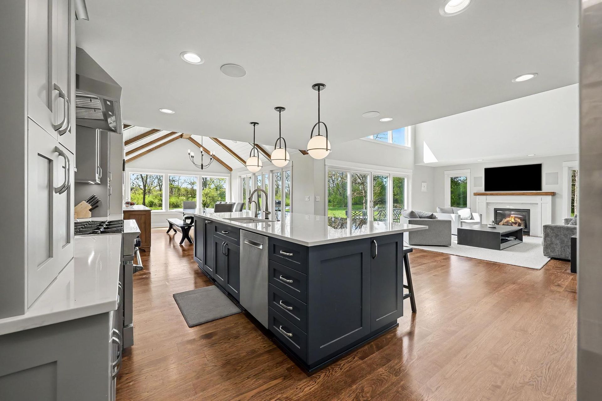 Remodeled open-plan kitchen with a navy island under globe pendants, hardwood floors, and a family room with a lit fireplace beyond.