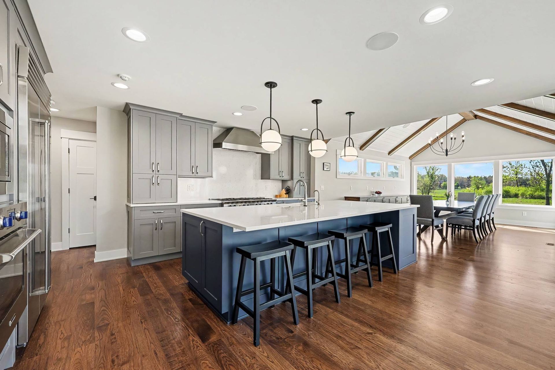 Remodeled kitchen with light gray shaker cabinets, a navy island with quartz countertops and four stools, and a 48-inch gas range with stainless hood.