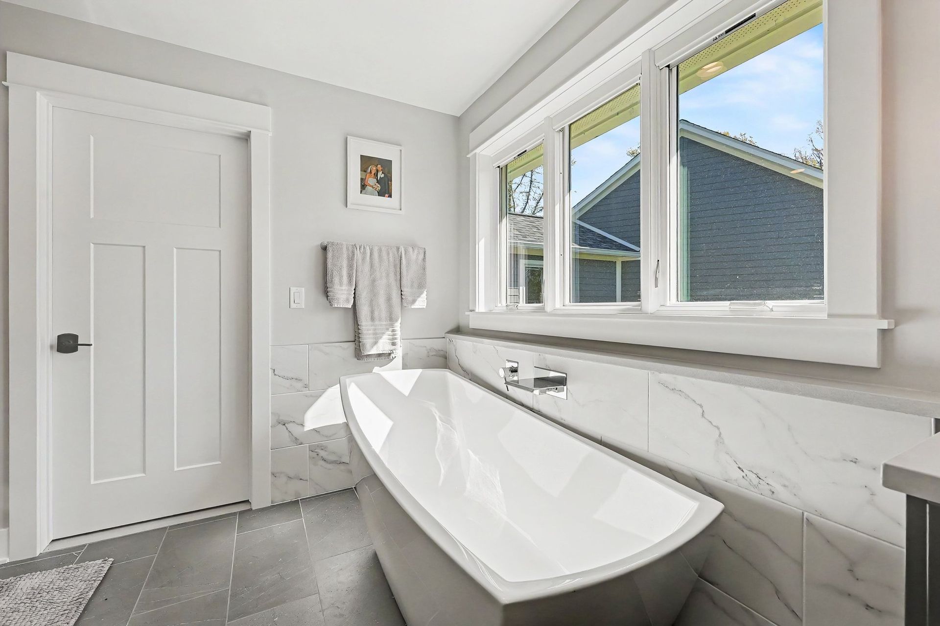 Primary bathroom with a freestanding soaking tub under casement windows, marble-look porcelain surround, quartz ledge, and large-format gray floor tile.
