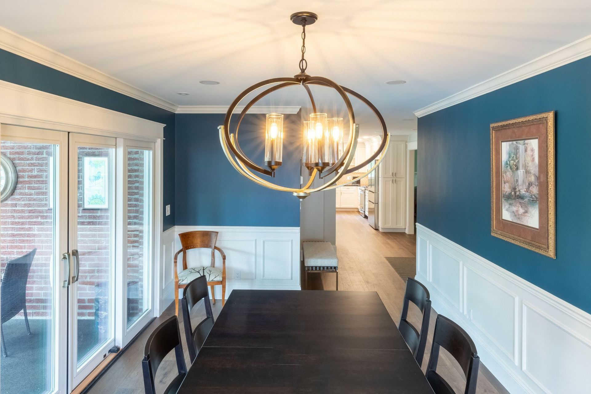 Dining table under an orb chandelier, with blue walls above white wainscoting and a view down the hall to the kitchen.