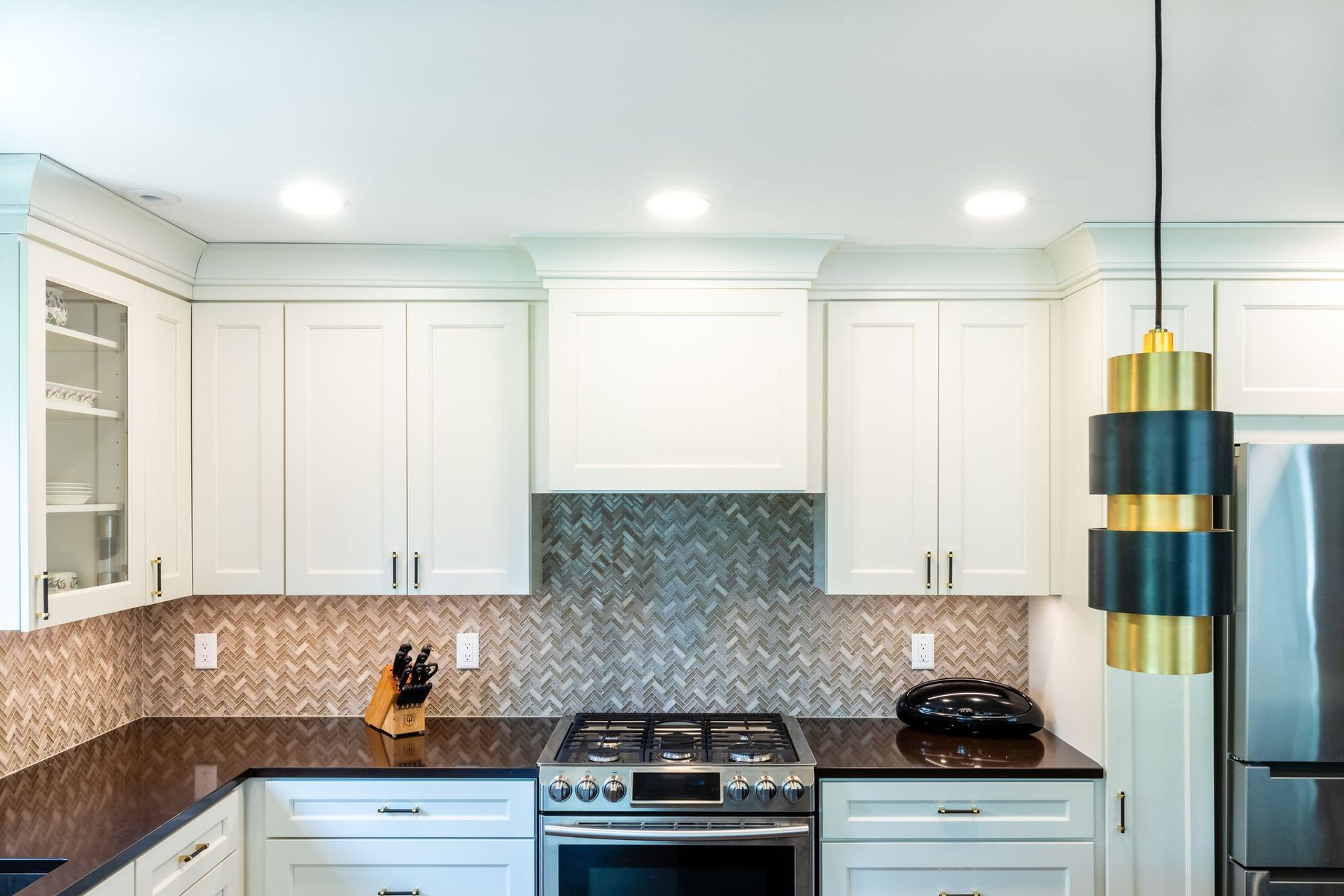 Kitchen range wall with a gas range, cabinetry range hood, herringbone mosaic backsplash, and dark countertops.