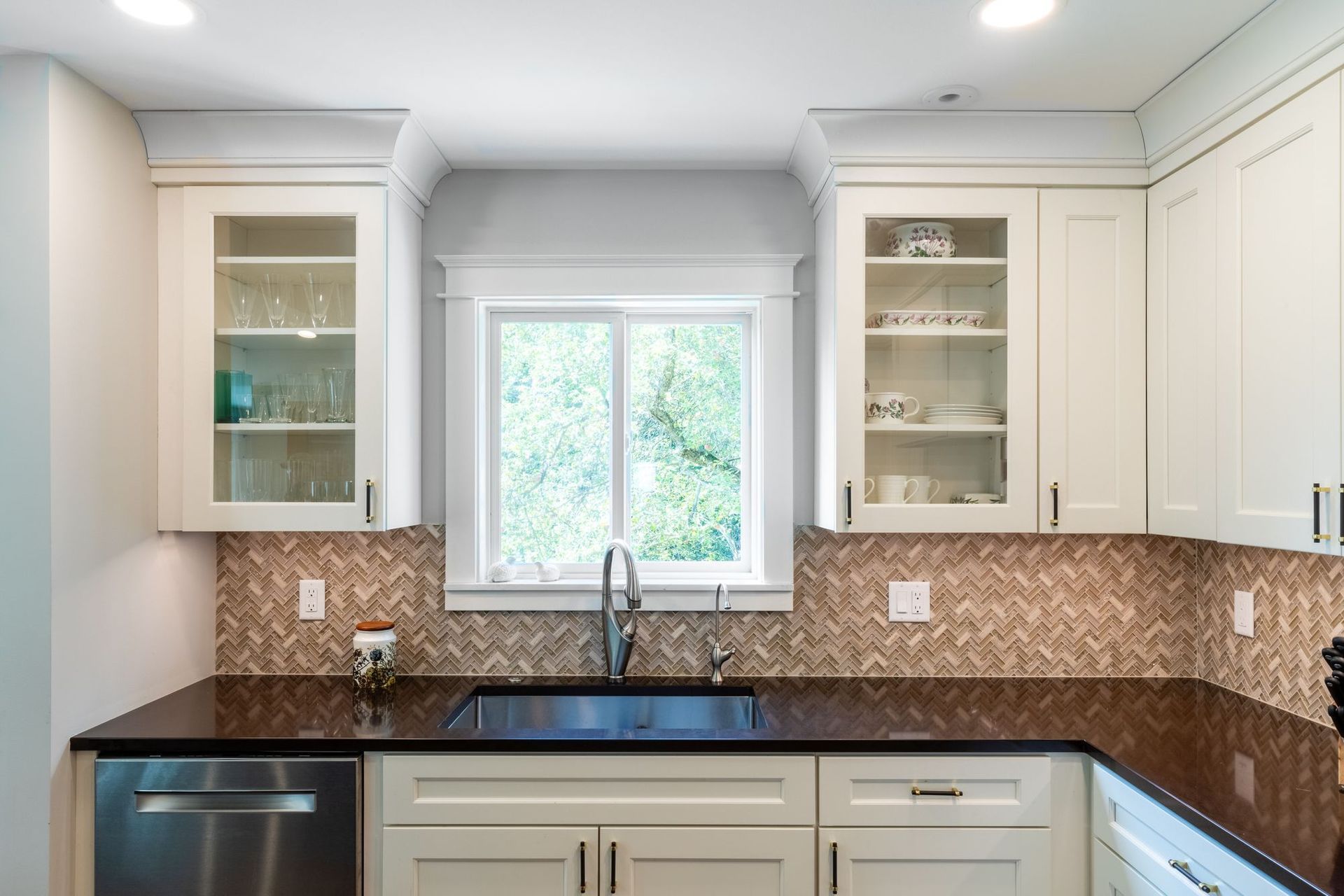Kitchen sink under a window framed by glass-front upper cabinets, with a herringbone tile backsplash.