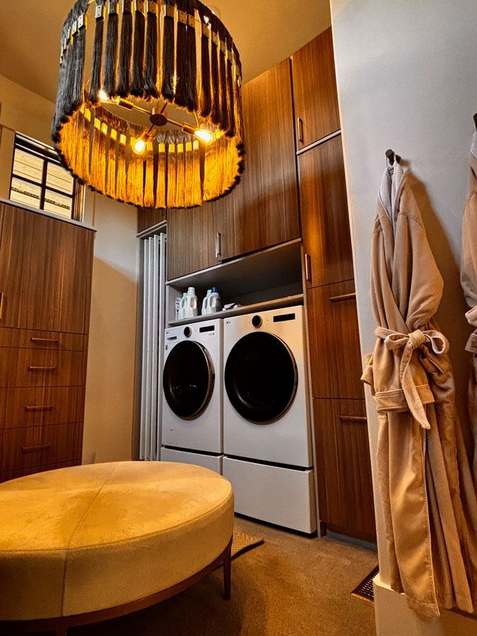 Laundry room with washer-dryer, wooden cabinets, round yellow ottoman, and hanging robe under a woven pendant light