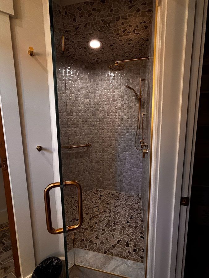 Small tiled shower stall with glass door and brass handle, lit by a ceiling light.