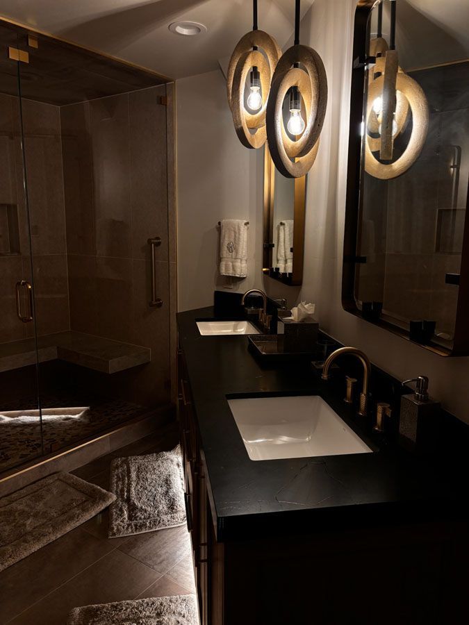 Dim restroom with black vanity, white sink, round lit mirrors, and a walk-in shower.