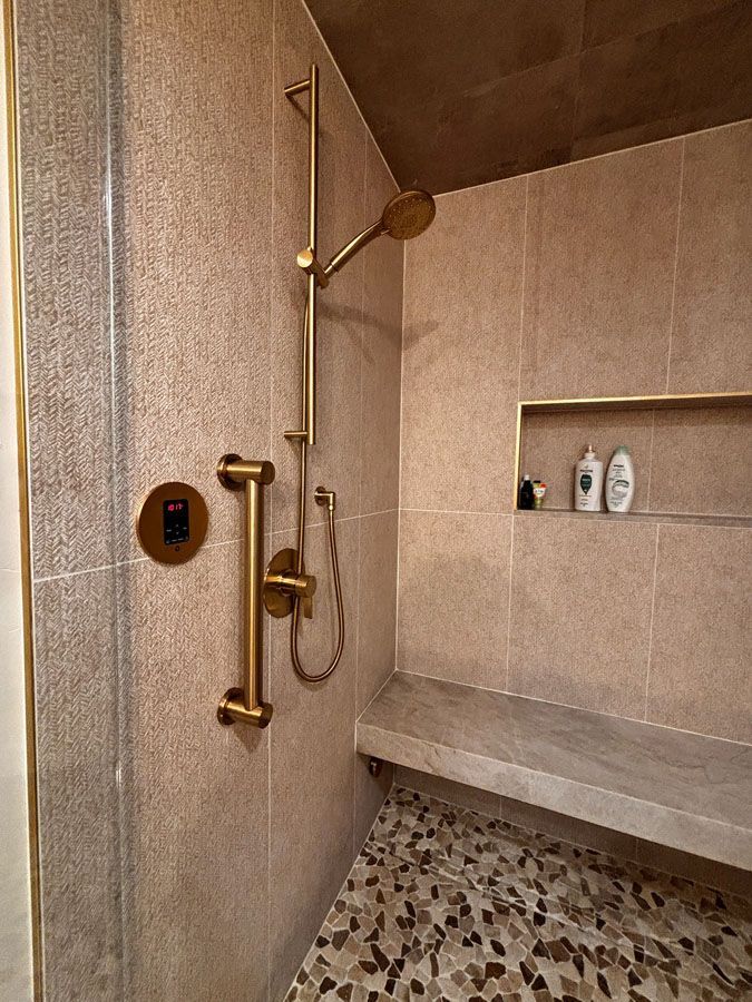 Accessible shower with gold grab bars, handheld showerhead, built-in shelf, and pebble-tile floor.