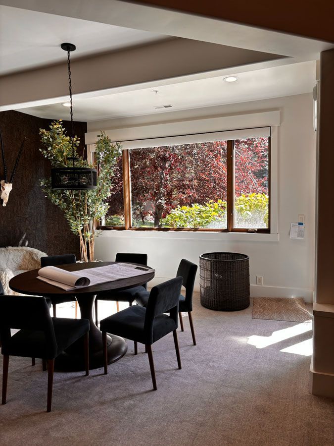 Dining room with round black table, chairs, and large window overlooking a sunlit garden.