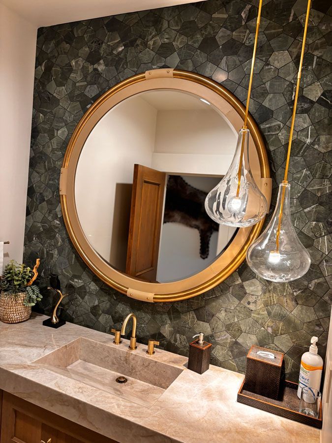 Modern bathroom vanity with round gold mirror, stone sink, hanging glass pendants, and dark textured wall.