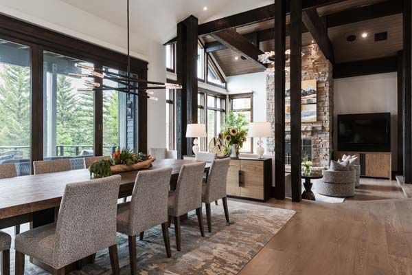 Modern open-plan dining and living room with wood beams, large windows, and a dining table with chairs.