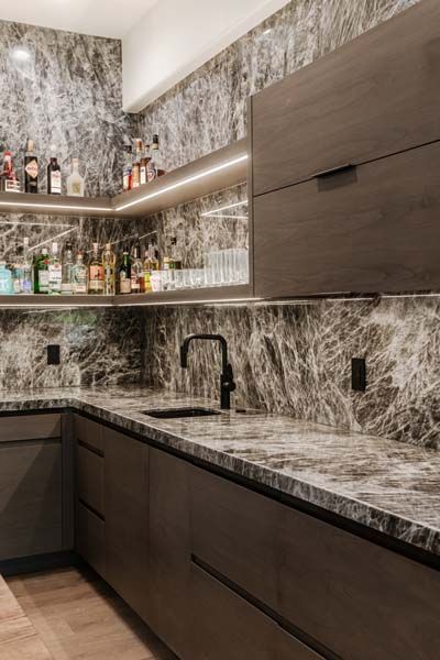 Modern kitchen bar with dark wood cabinets, marble-patterned counters and backsplash, and lit liquor shelves