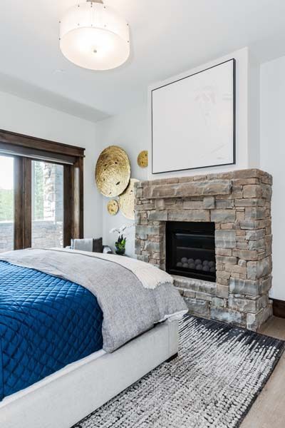 Bedroom with blue quilted bedspread, stone fireplace, and a window with wood trim