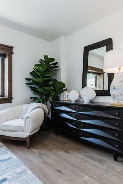 Modern bedroom corner with black dresser, mirror, white chaise lounge, and potted plant.