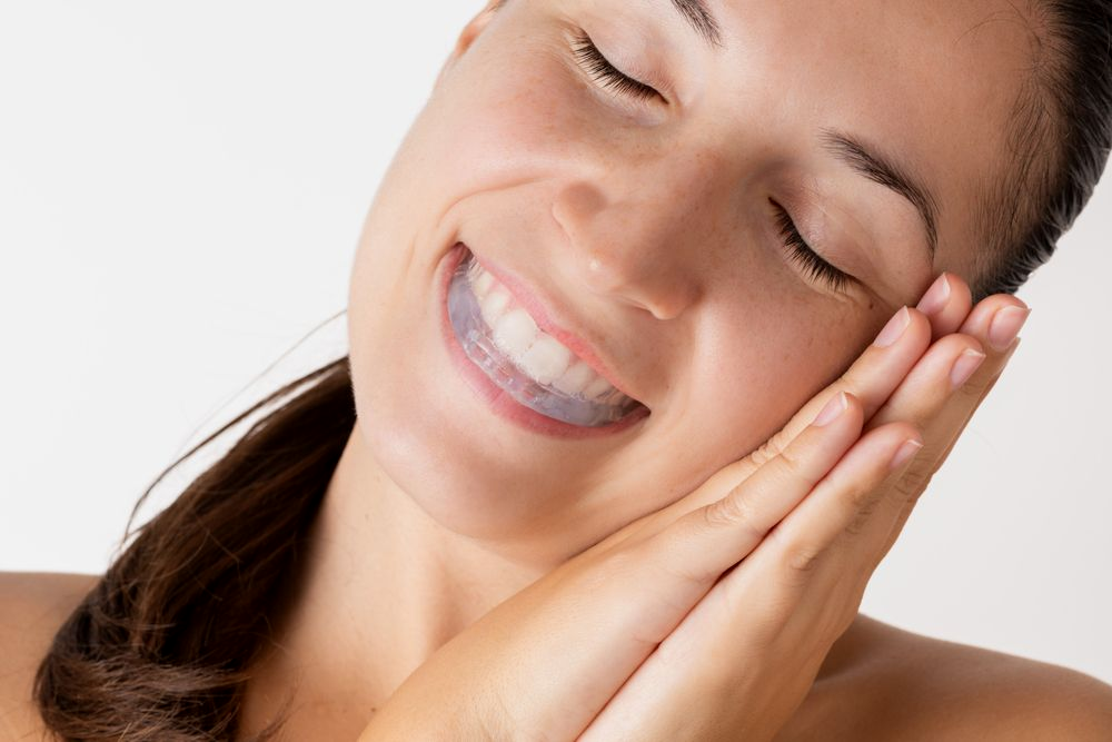Woman with closed eyes smiling, wearing a teeth whitening tray. Hands rest near face.