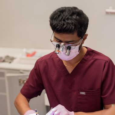 Dentist in blue scrubs, pink mask, and gloves working on a patient in a dental chair under bright light.