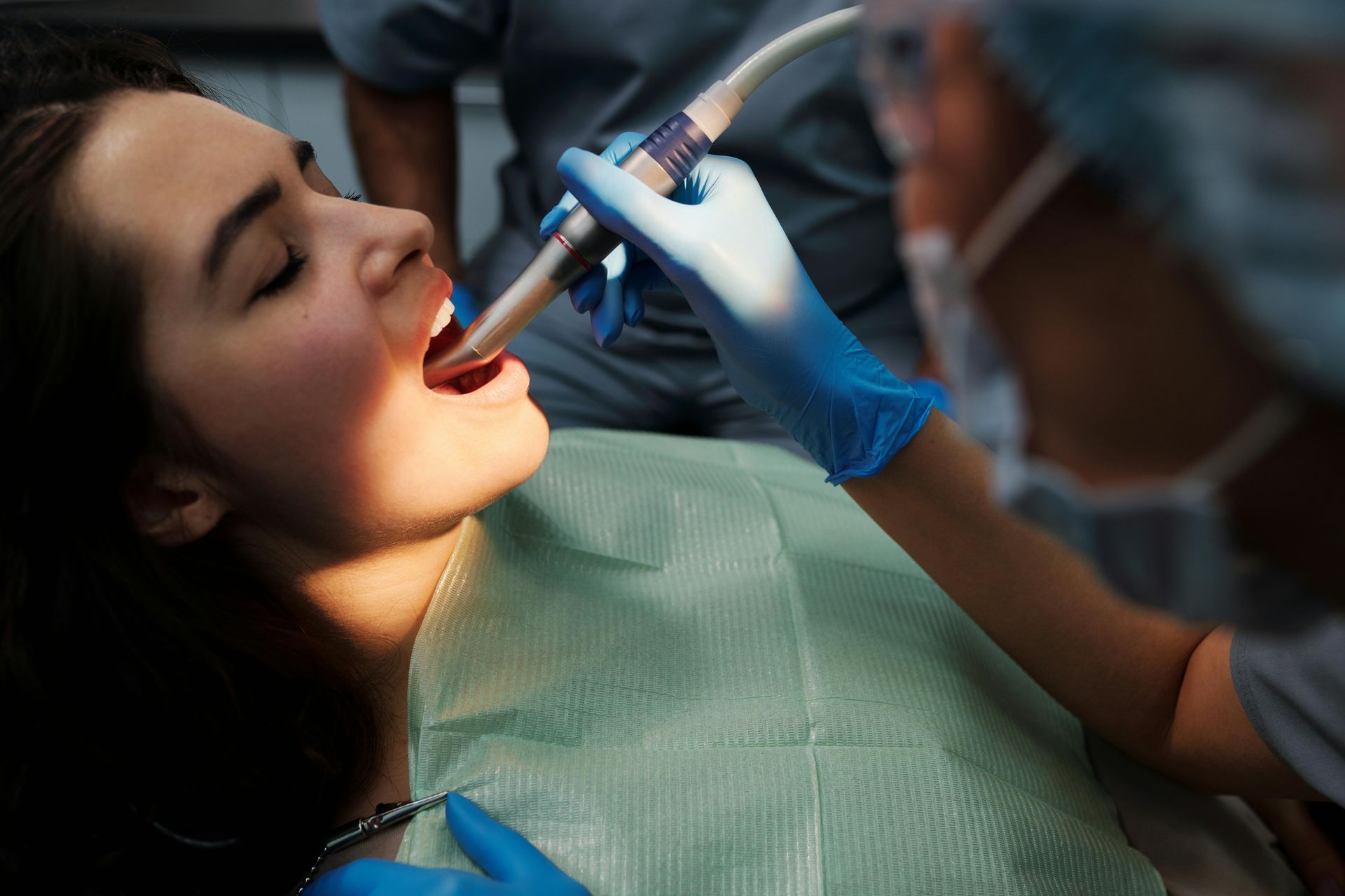 Woman in dental chair, mouth open while dentist uses a tool; blue gloves and surgical mask.