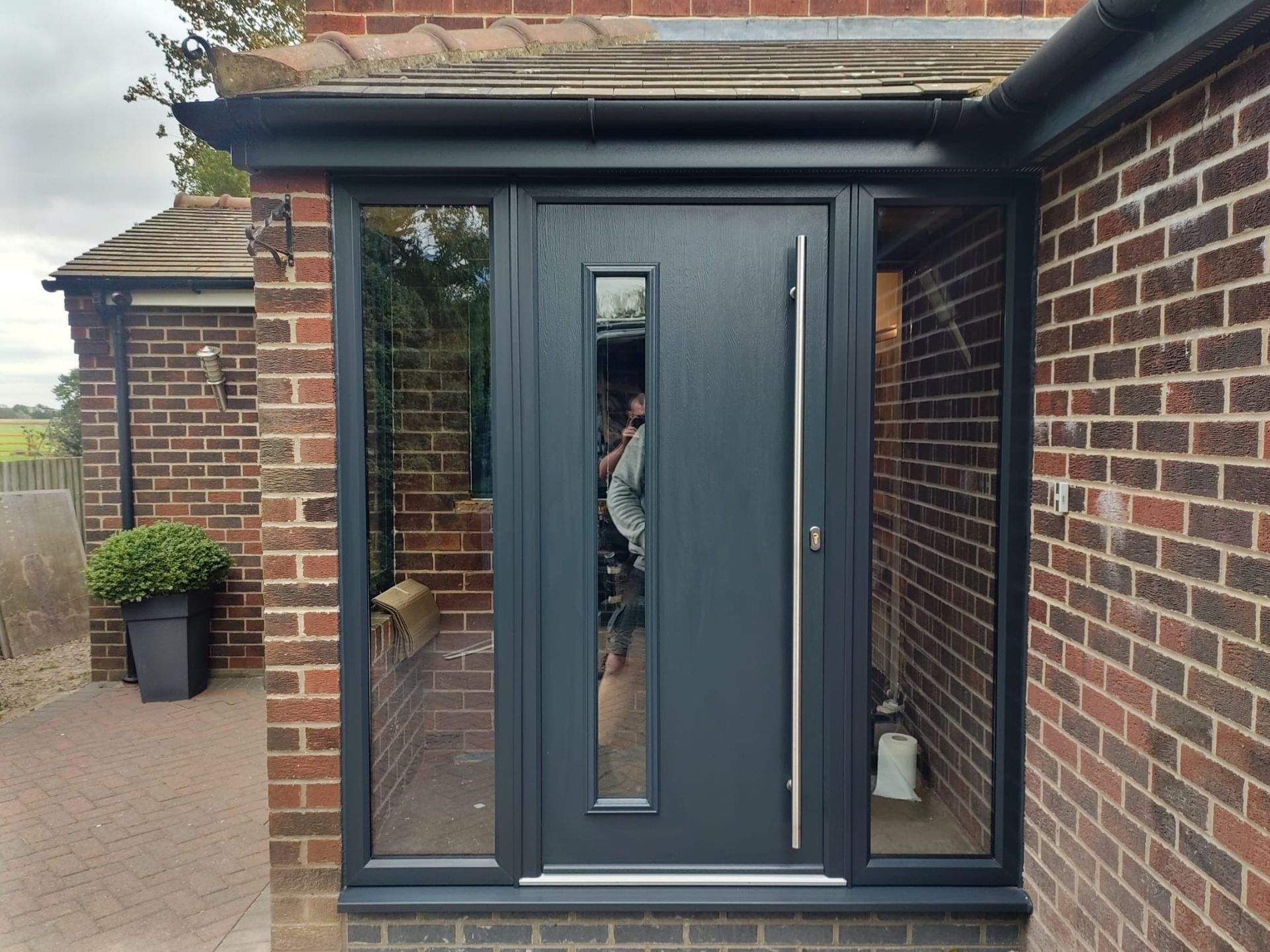 Brick house entrance with dark gray front door, narrow glass panels, and a brick patio path