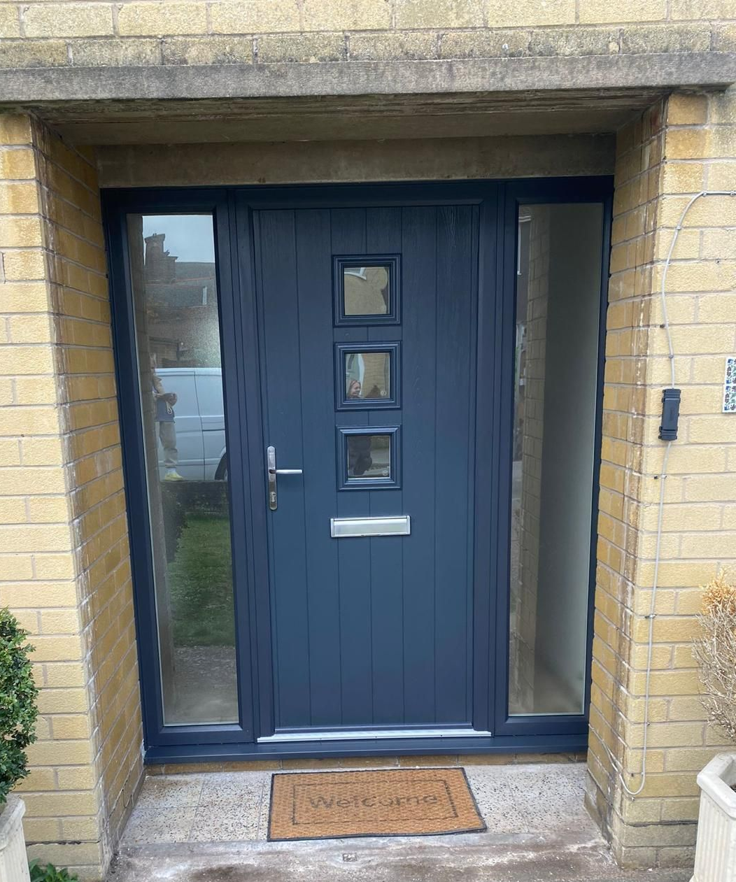 Dark blue front door with narrow side windows set in a brick entryway, with a doormat outside.