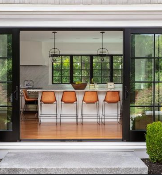 Modern kitchen with orange bar stools, black-framed glass doors, and pendant lights opens to a patio.