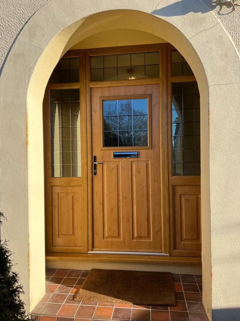 Light oak front door with side panels, a square glass window, and a silver letterbox, set in a stone-arched entryway.