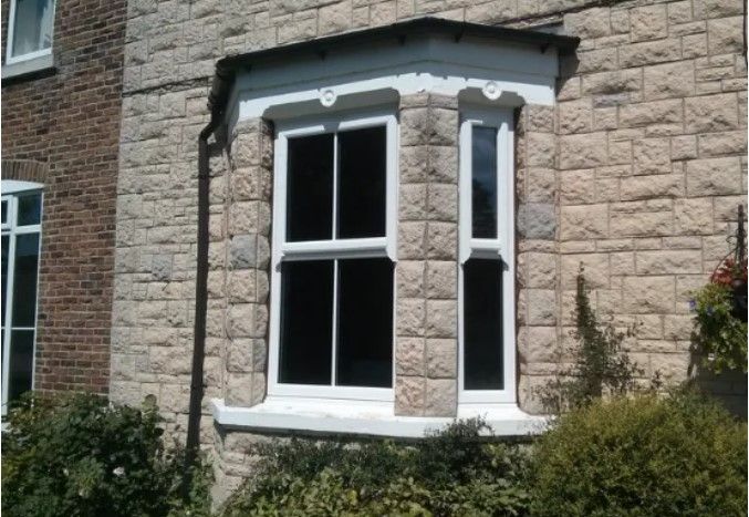 Stone house exterior with a white bay window and dark trim, framed by shrubs.