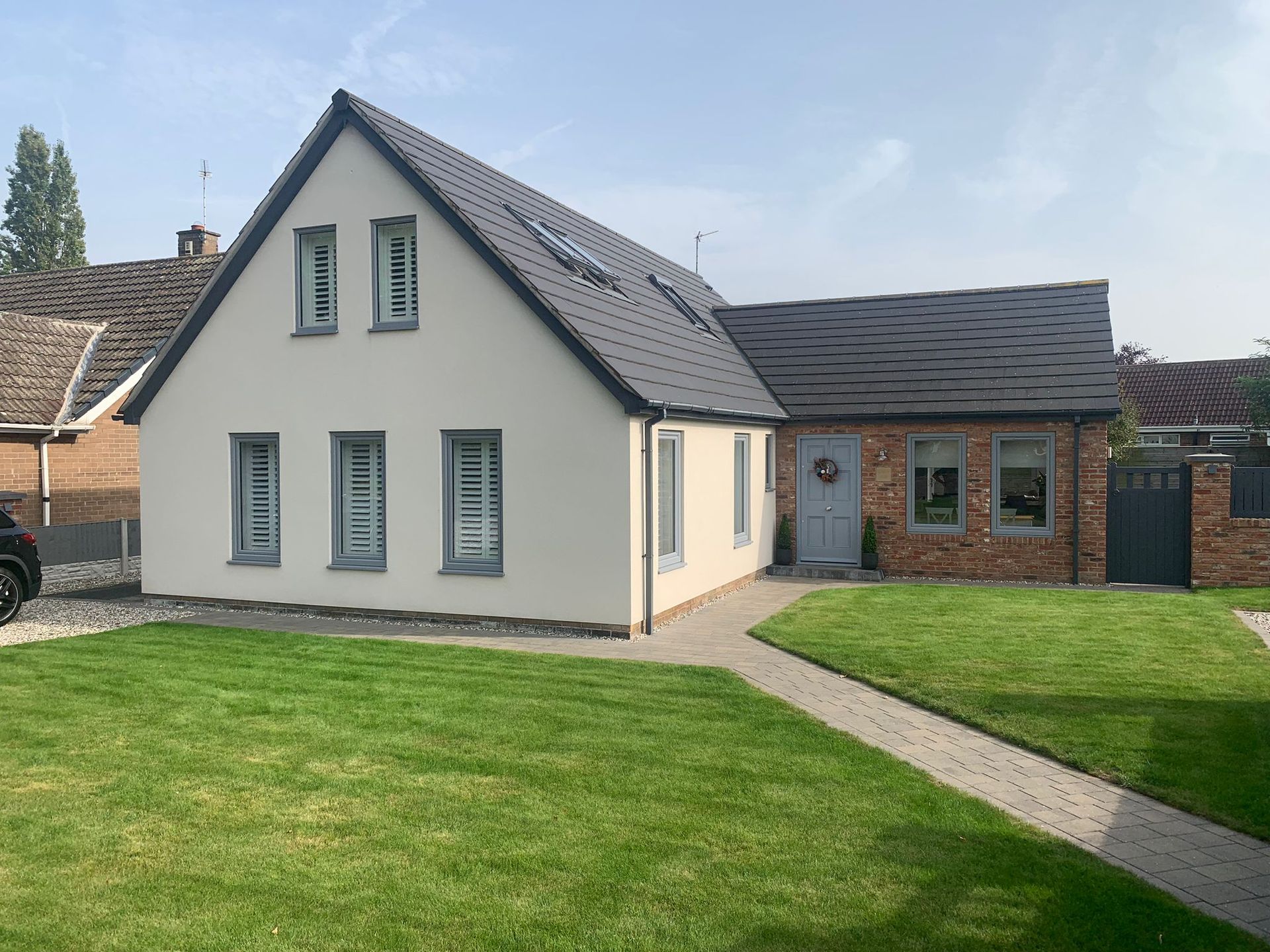 Modern house with gray roof, white walls, brick side extension, and a green lawn under a cloudy sky