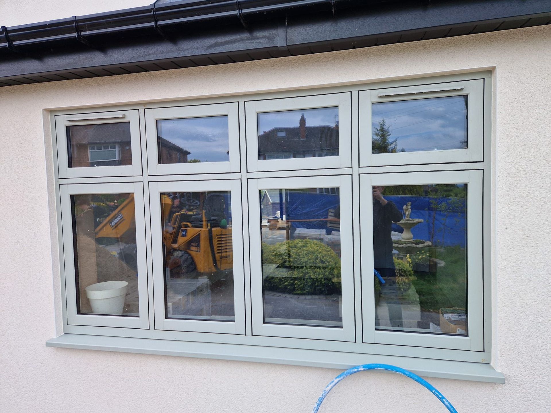 White-framed window reflecting a blue excavator and greenery, with a small white plant pot inside.