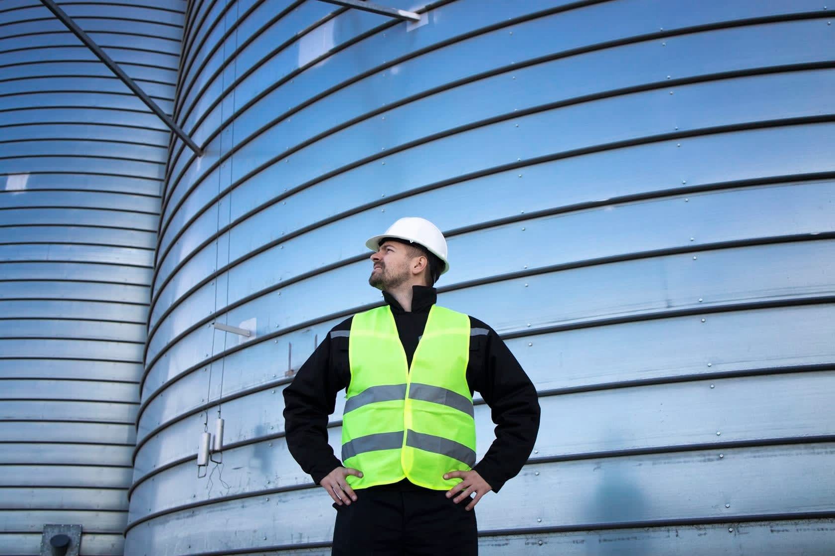 Worker in a white hard hat and neon safety vest standing before large industrial tanks