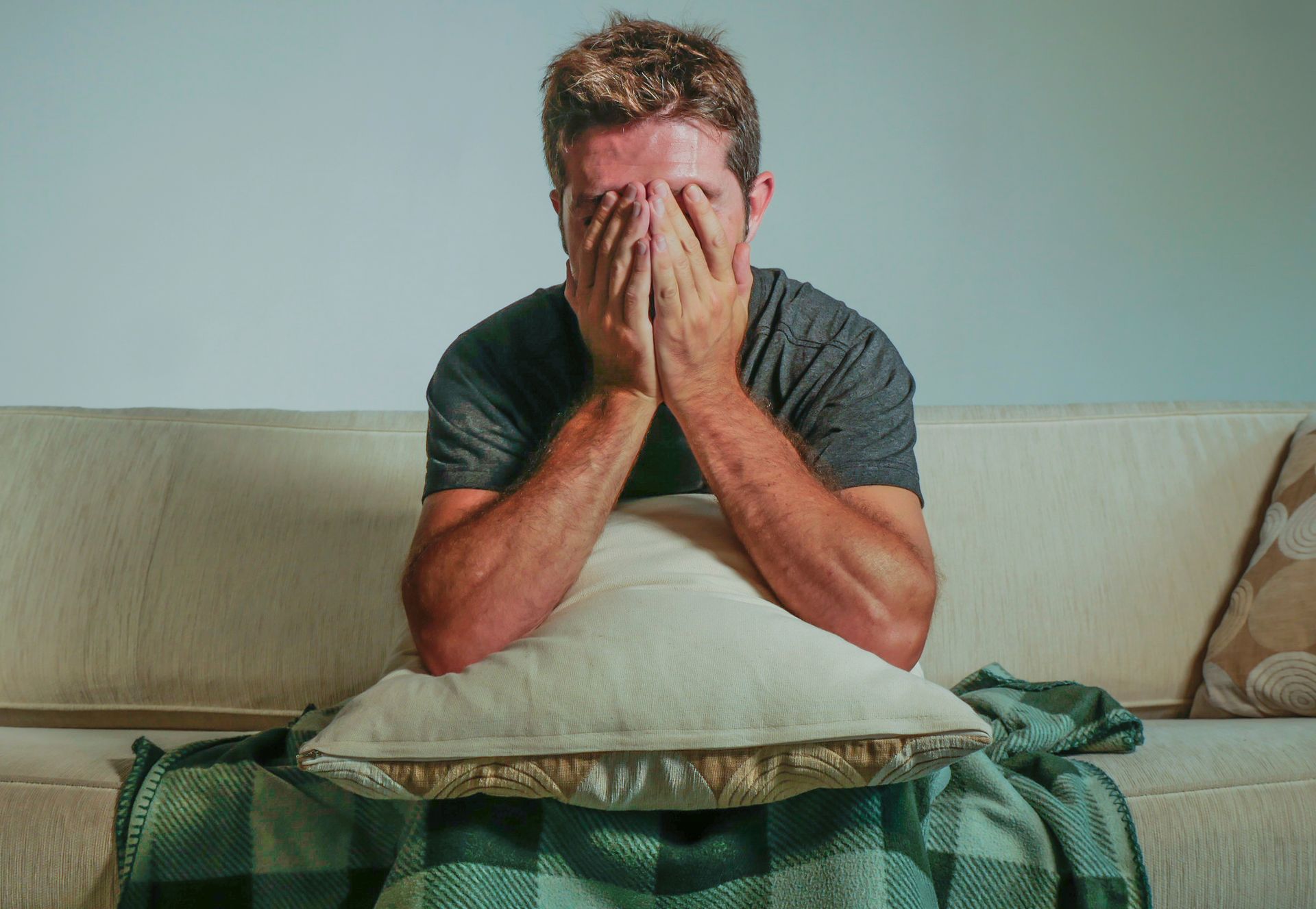 Man sitting on a couch, covering his face with his hands, pillow in lap, blanket, distressed expression.
