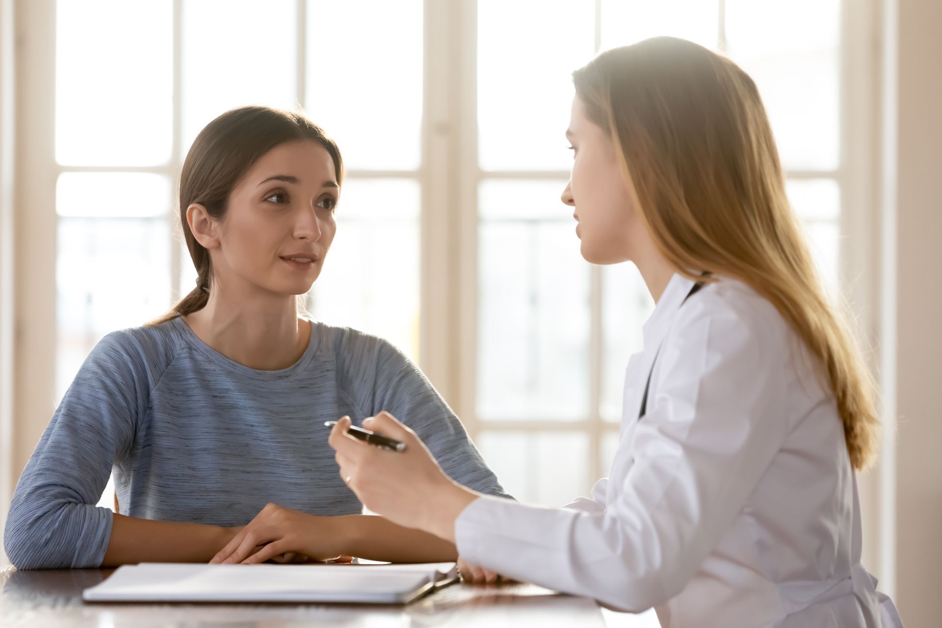 Woman in blue shirt speaking with a doctor in a white coat at a desk. Sunlight streams from a window.