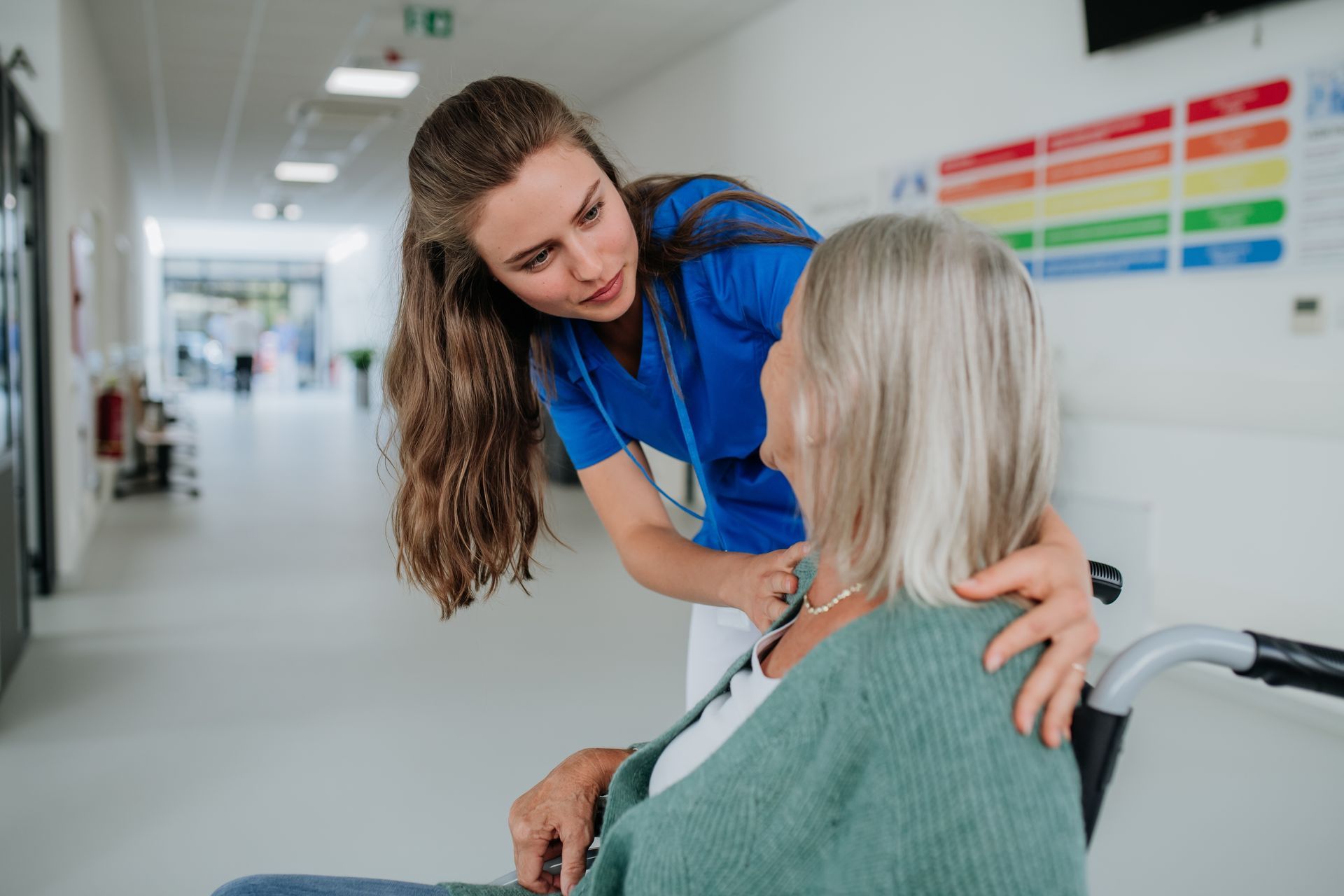 Nurse in blue scrubs comforts patient in wheelchair in hospital hallway.