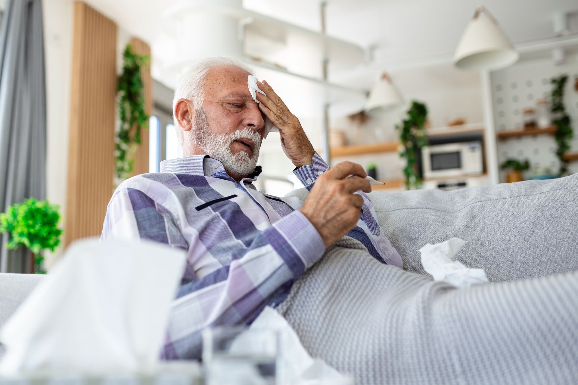 Man with a thermometer in hand, touches his forehead with a cloth, seated on a couch with tissues.