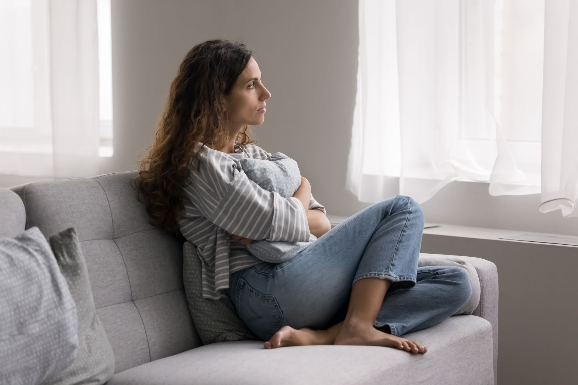 Woman sitting on couch, looking out window, hugging a pillow, contemplative expression.