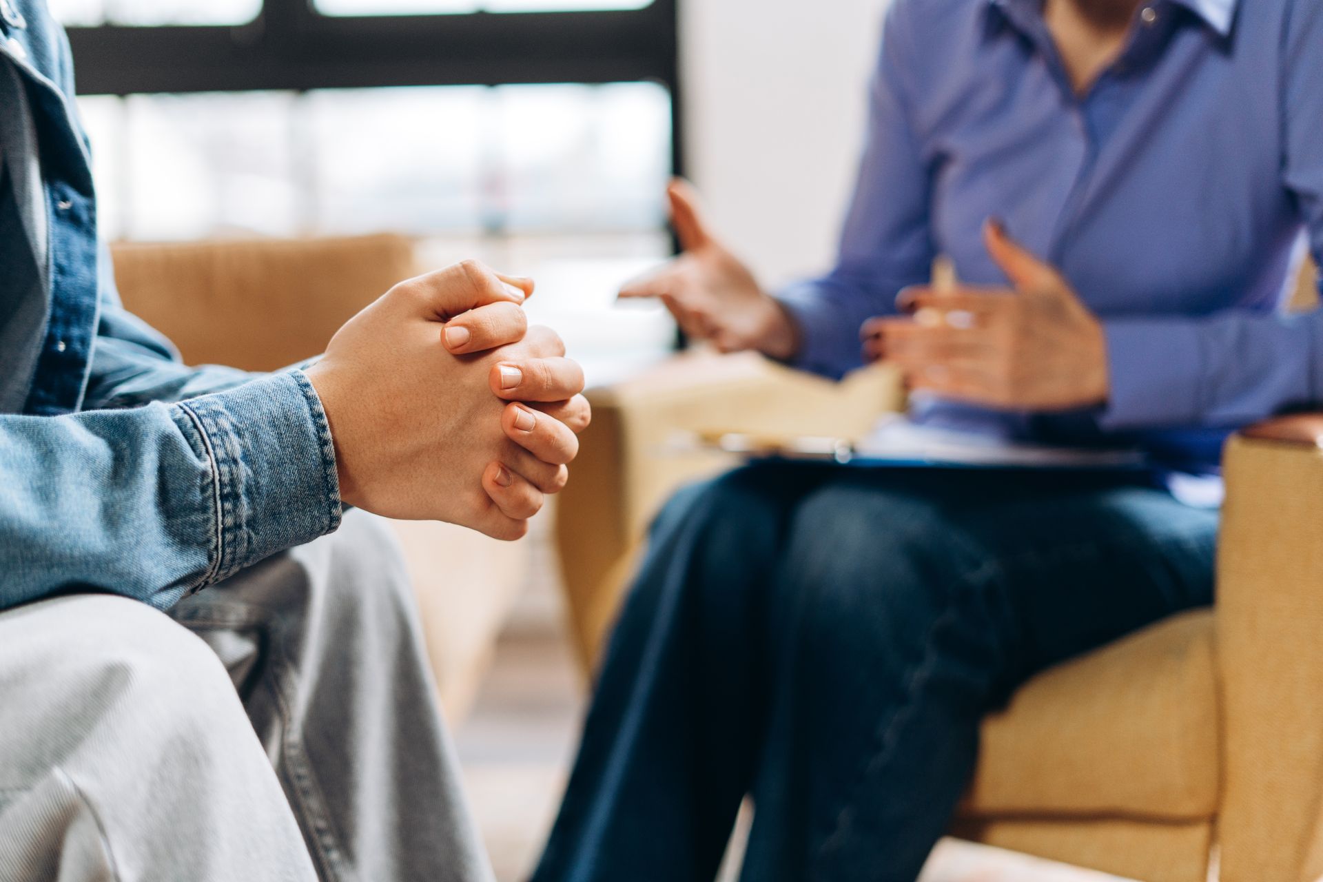 Person in denim jacket with hands clasped, facing a person in blue shirt gesturing, in a counseling session.