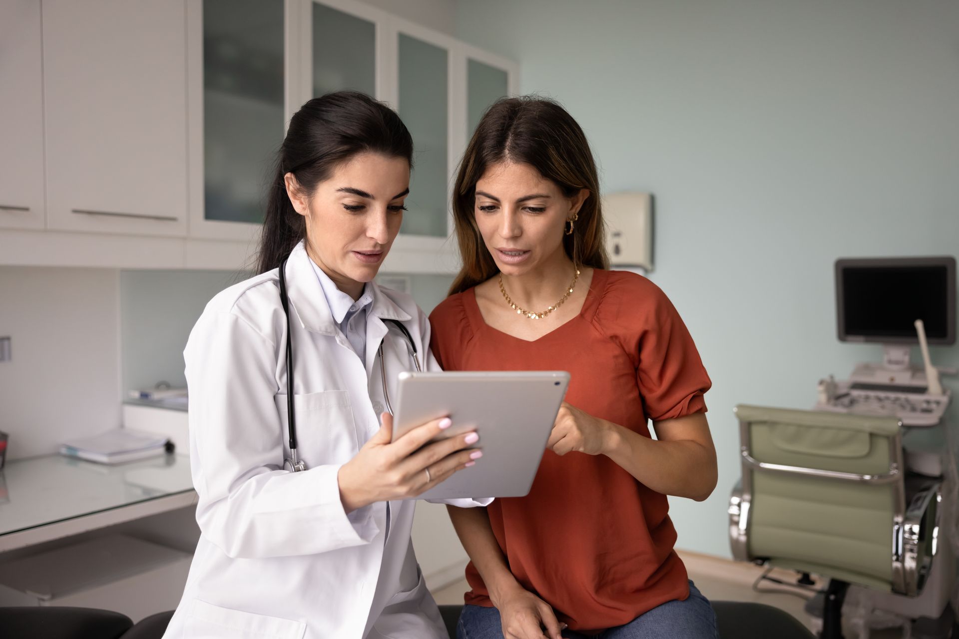 Doctor in white coat holds clipboard, consulting with a patient in a beige cardigan in a medical office.