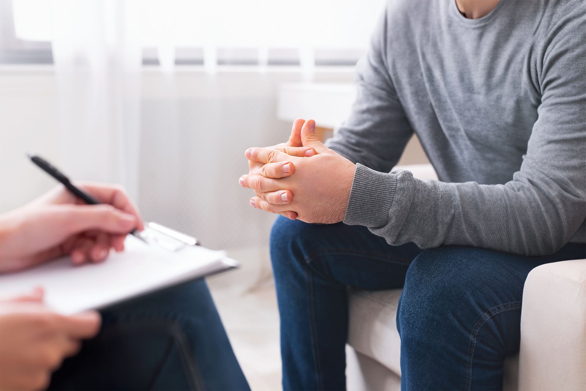 Therapist taking notes while speaking with a seated client in a bright office.