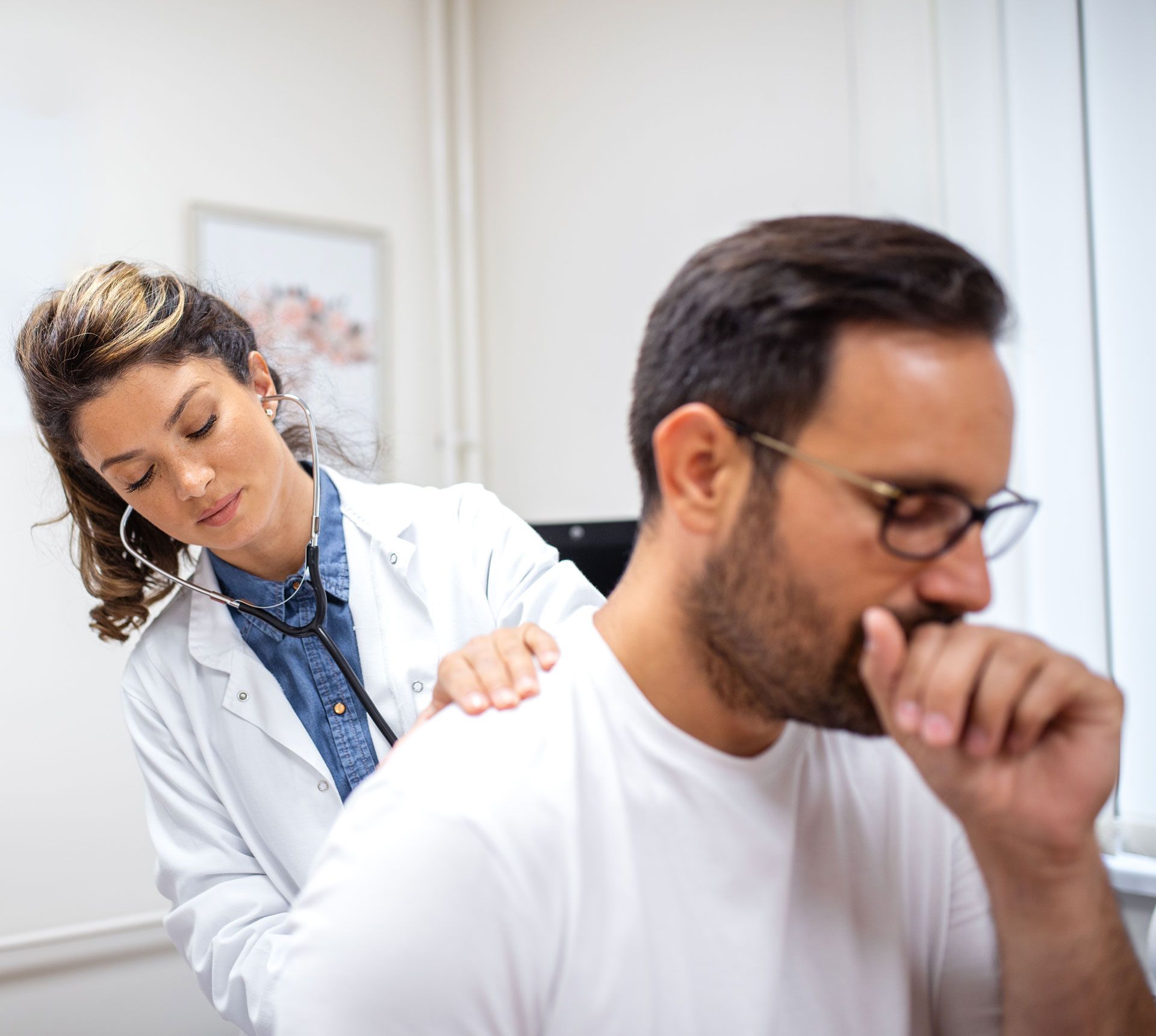 Doctor examining a seated patient in a bright clinic, with the patient coughing into his hand.