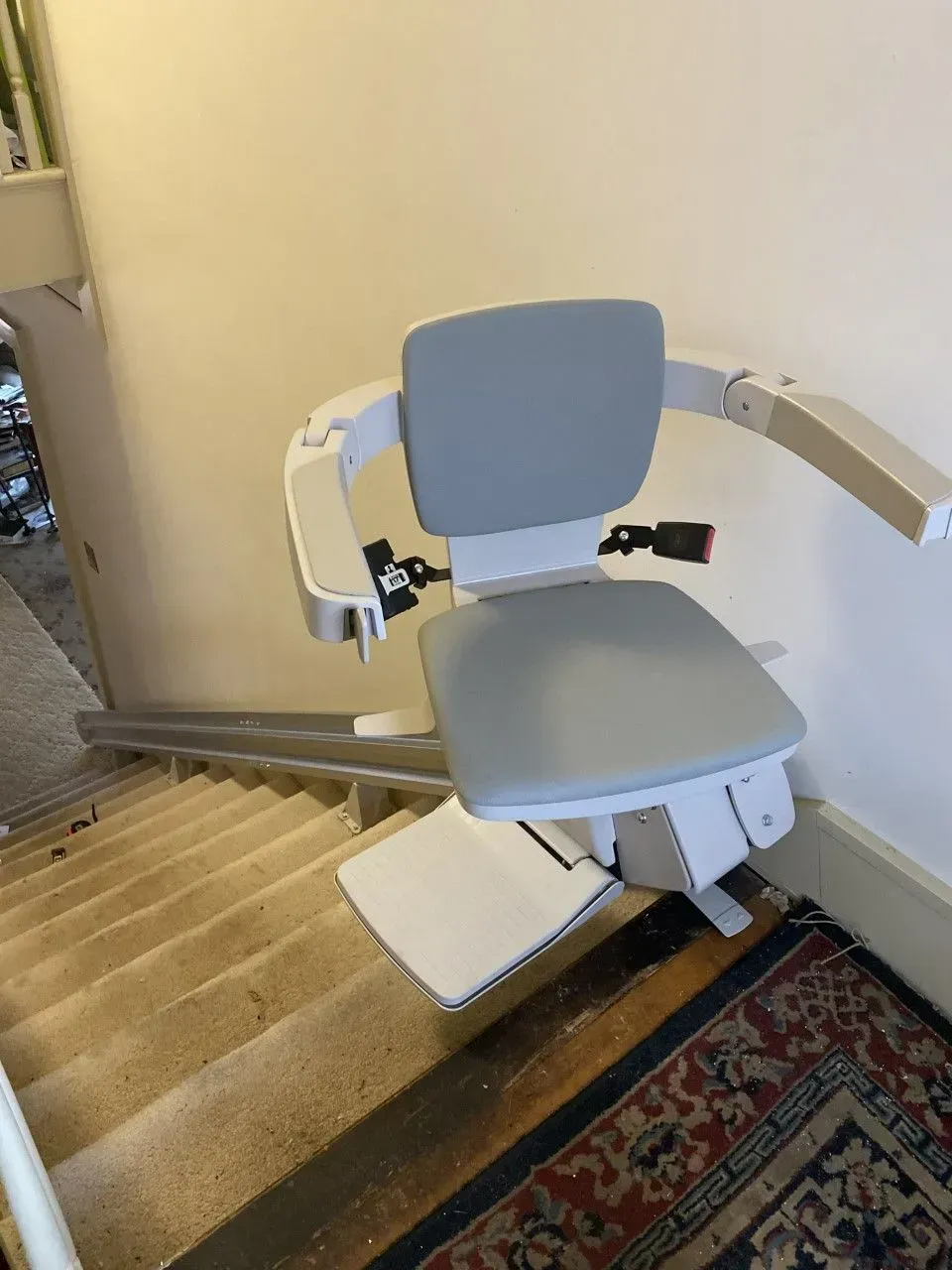 Stairlift chair at the top of a carpeted staircase beside a wall and rug
