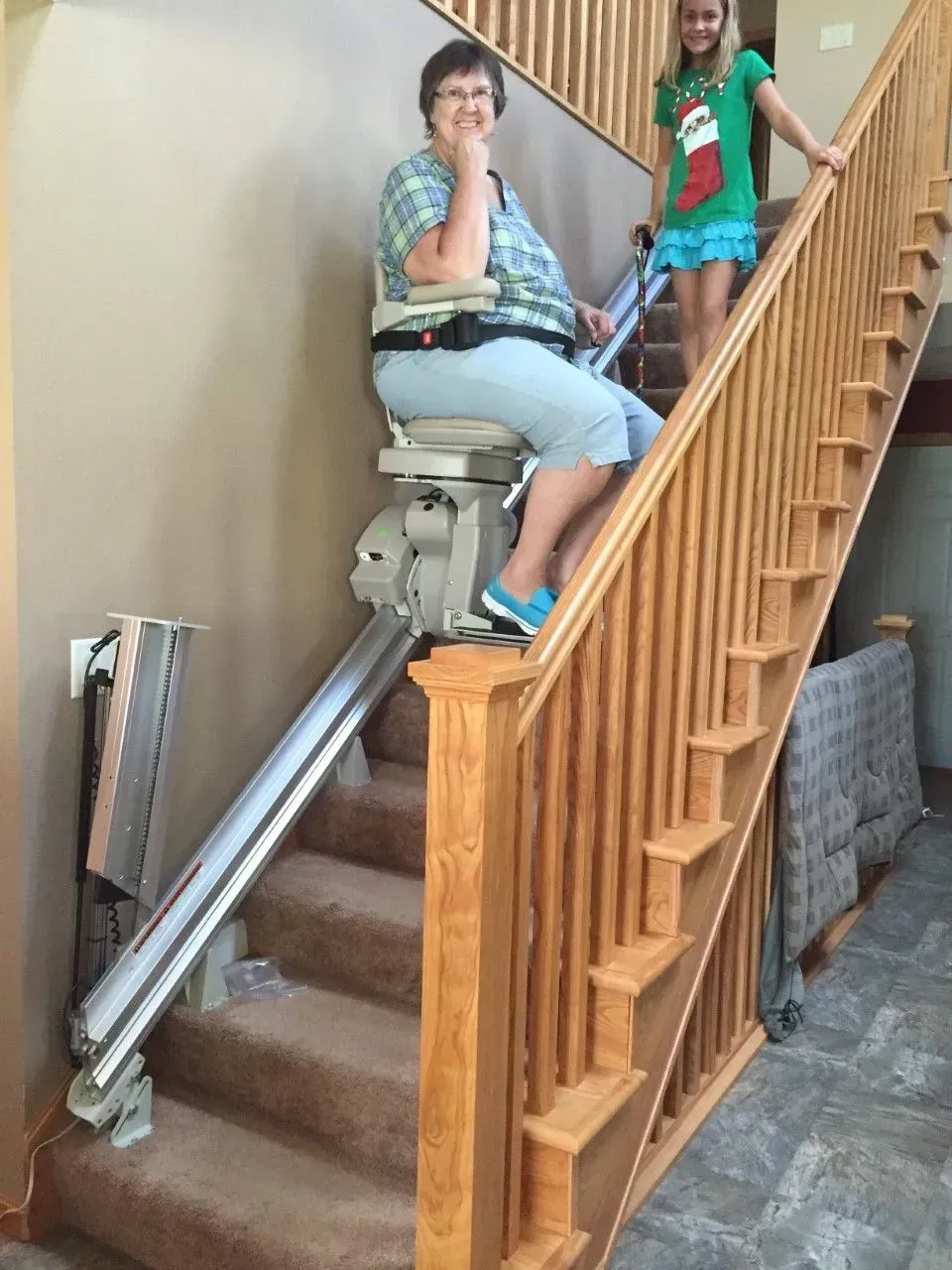 Woman rides a stair lift beside a wooden staircase, with another person standing nearby.