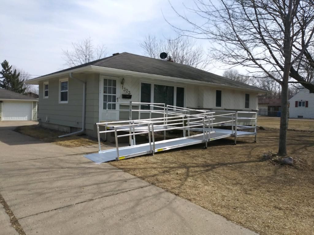 Single-story house with a white wheelchair ramp leading to the front door, beside a driveway and leafless tree.