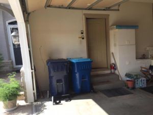 Garage entry with two blue trash bins, steps to a front door, and potted plants nearby