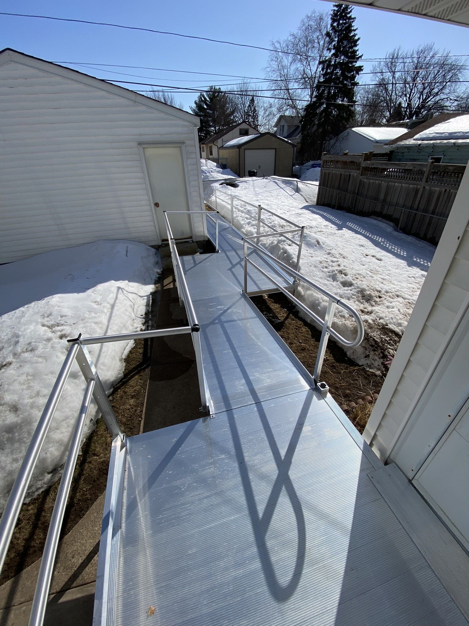 Snowy backyard walkway beside a white house, with metal railings and blue sky.
