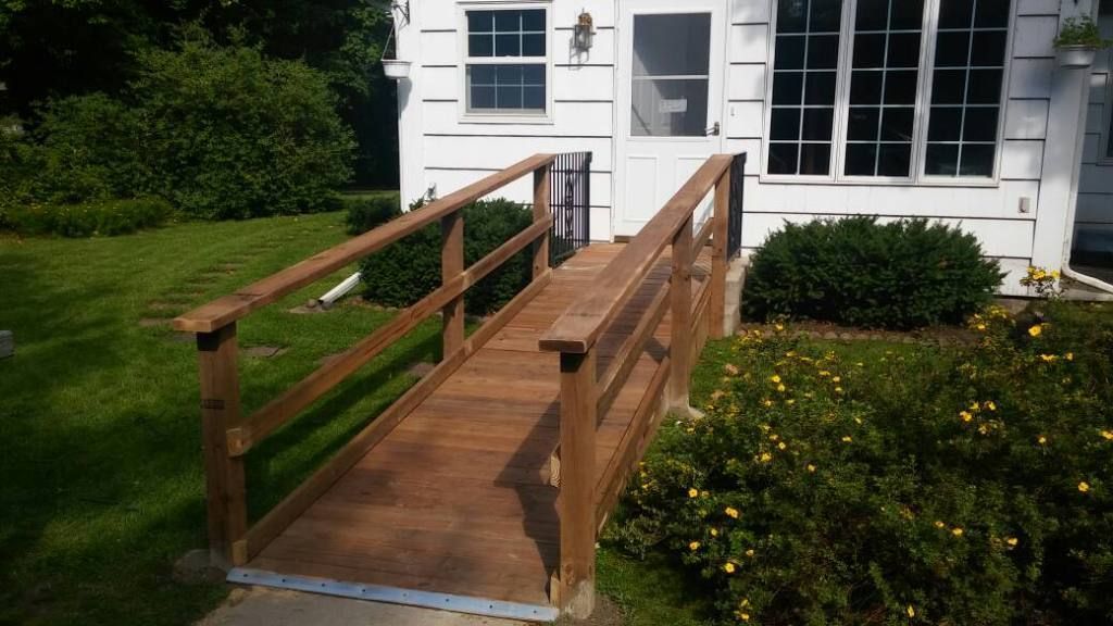 Wooden accessibility ramp leading to a white house entrance with steps and railings, beside a garden.
