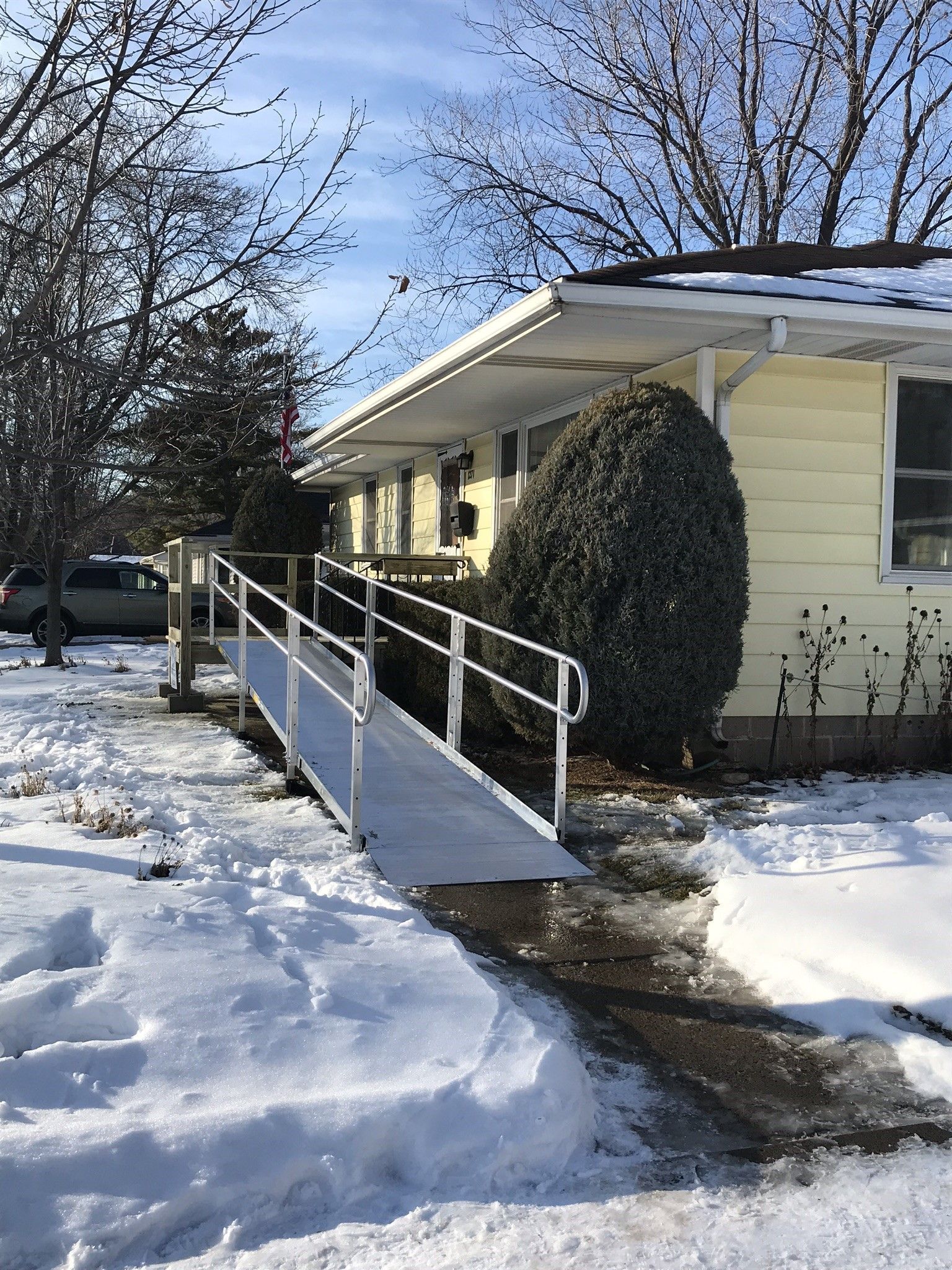 Snowy front walkway with a wheelchair ramp leading to a yellow house under bare trees
