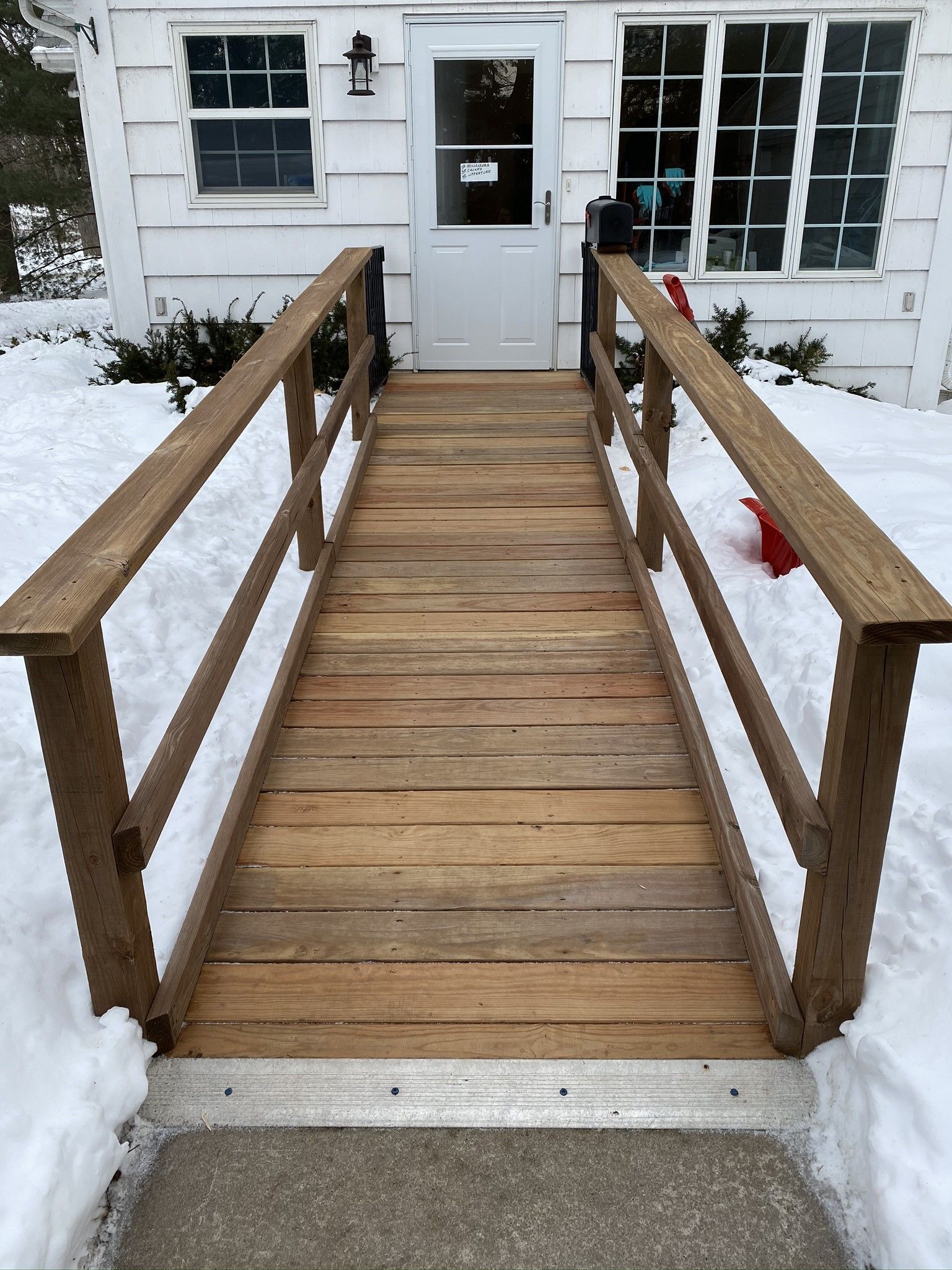 Wooden ramp with railings leading to a white house door, bordered by snow.