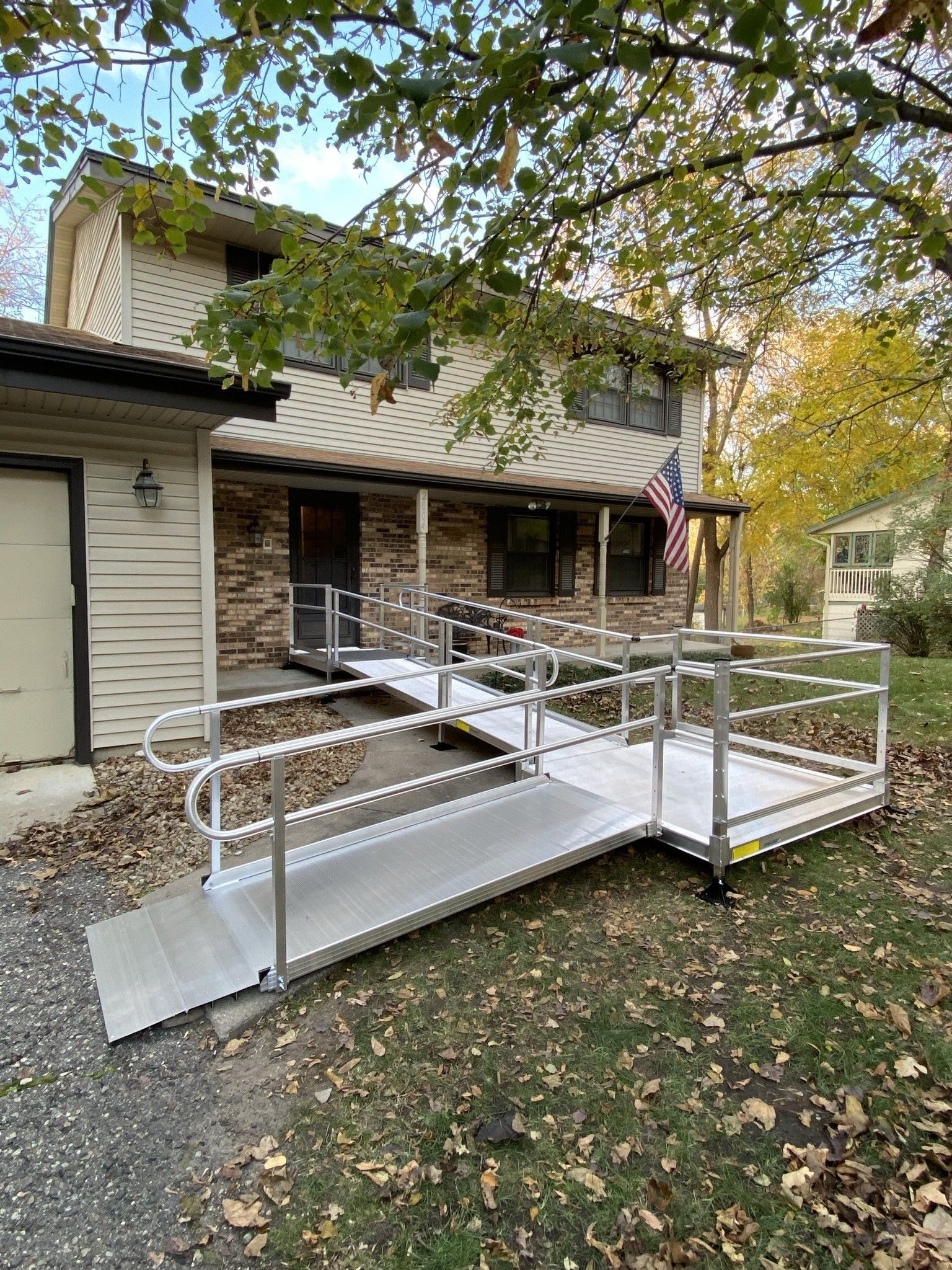 Modular aluminum ramp with railings leading to a house porch in a leafy yard.
