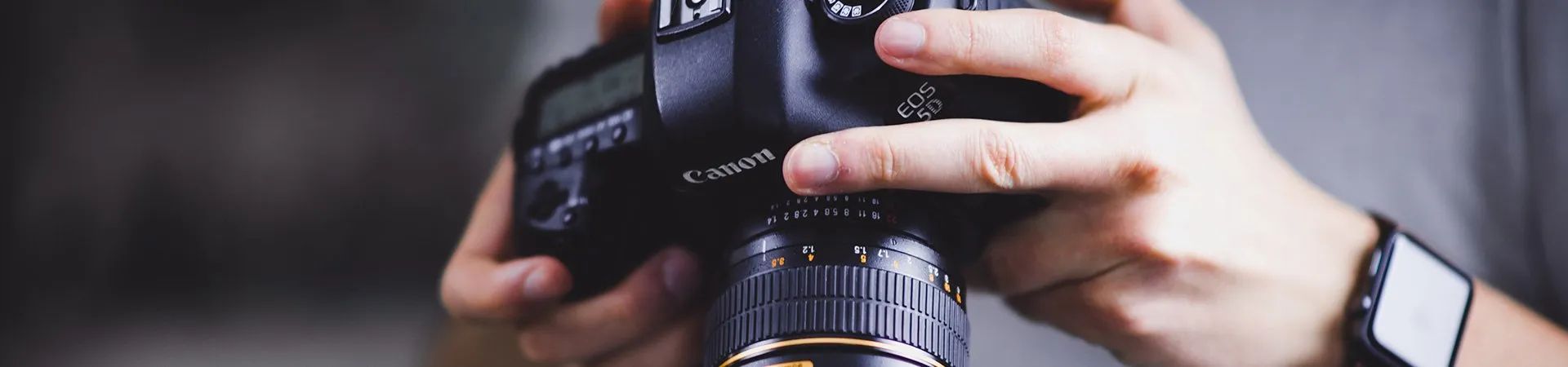 Hands holding a camera with a large lens, wearing a black wristwatch.